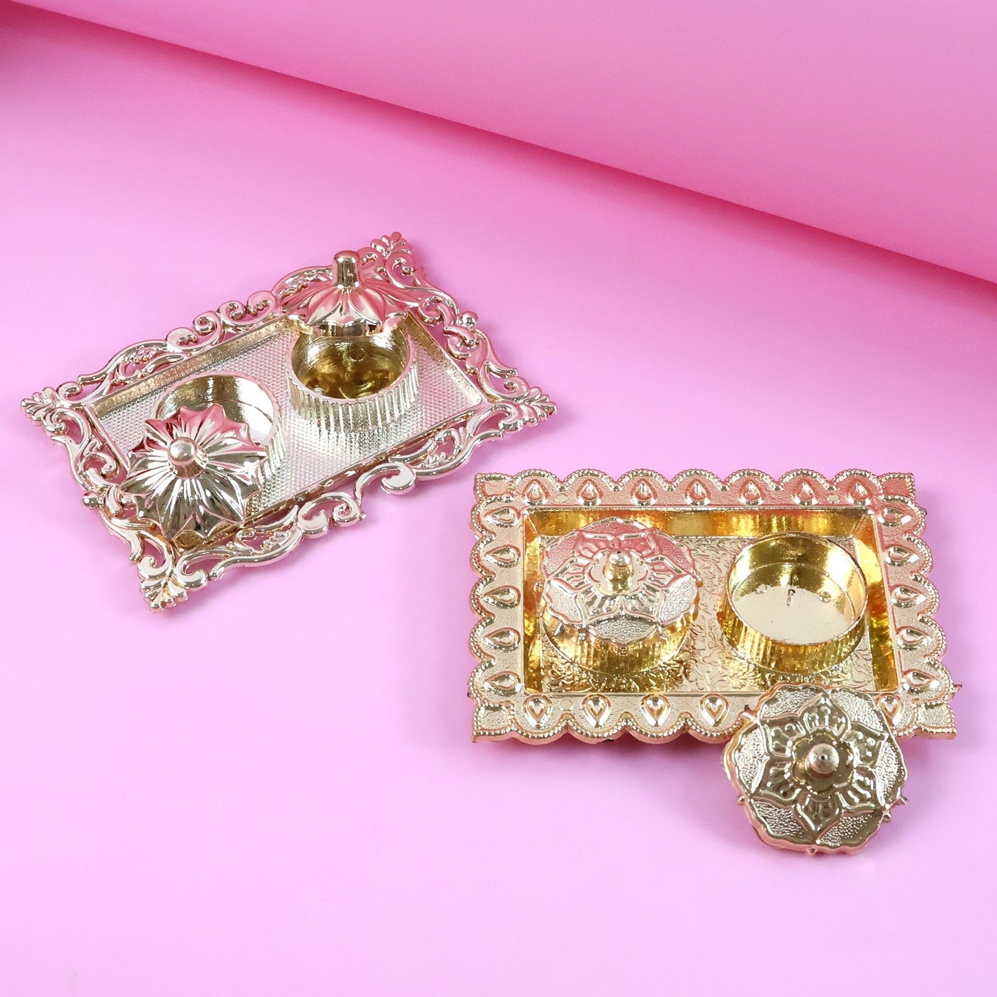 Two ornate rectangular pooja thalis, one gold and one silver, with small containers, on a pink background.