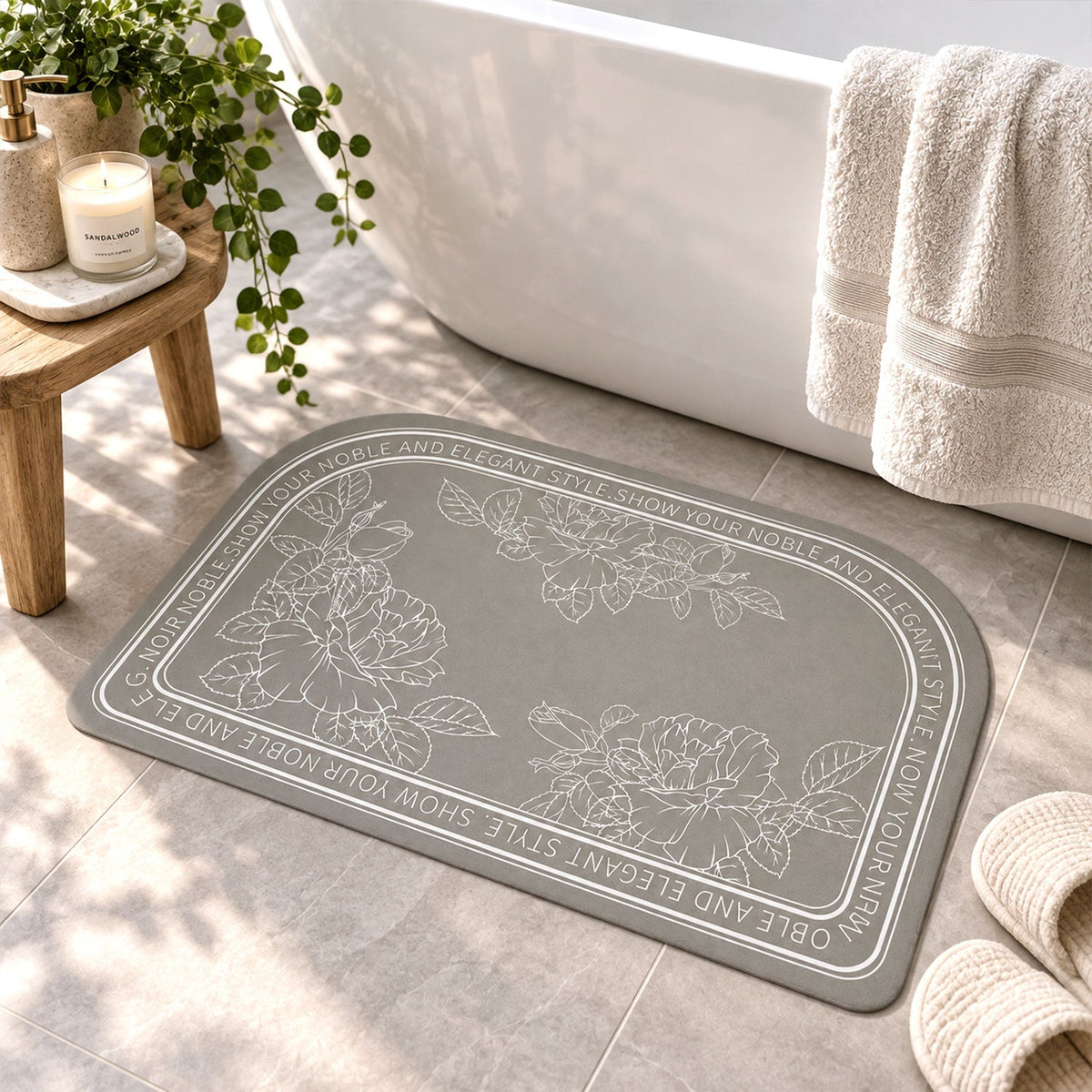 A gray half-round bath mat with white floral designs and text sits on a tiled floor next to a white bathtub.