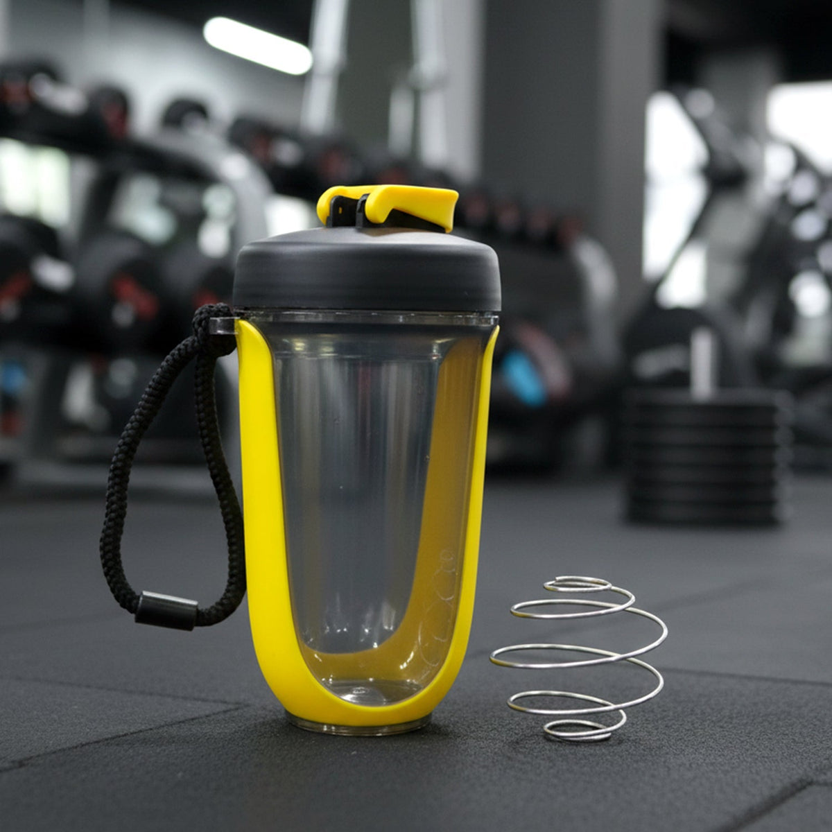 A yellow and black shaker bottle with a metal whisk sits on a dark gym floor with weights in the background.