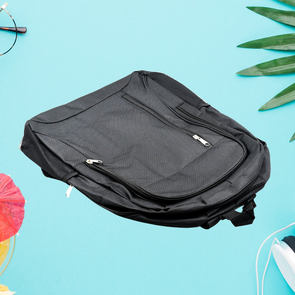 A black fabric backpack with multiple zippered pockets lies flat on a light blue surface with green leaves and round glasses