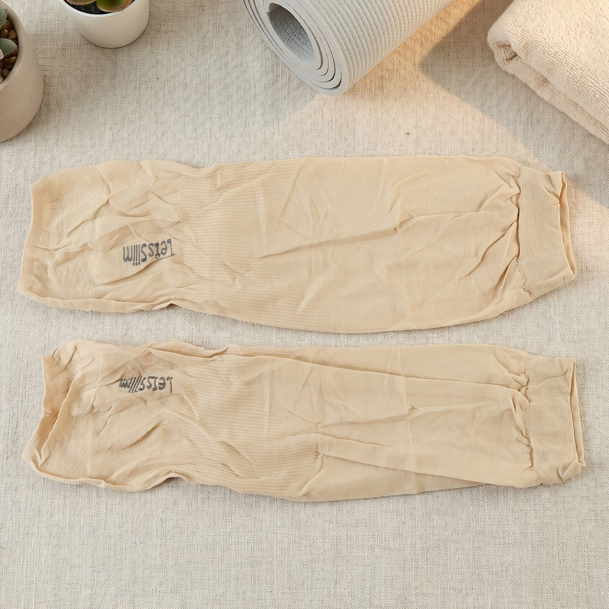 Two beige arm sleeves with "Let's Slim" logo, laid flat on a textured surface with a yoga mat and towel.