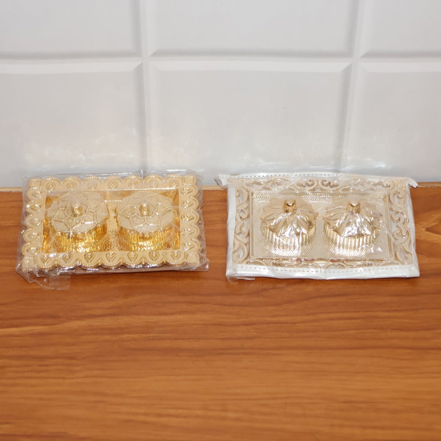 Two ornate rectangular pooja thalis, one gold and one silver, both in clear plastic packaging, on a wooden surface.