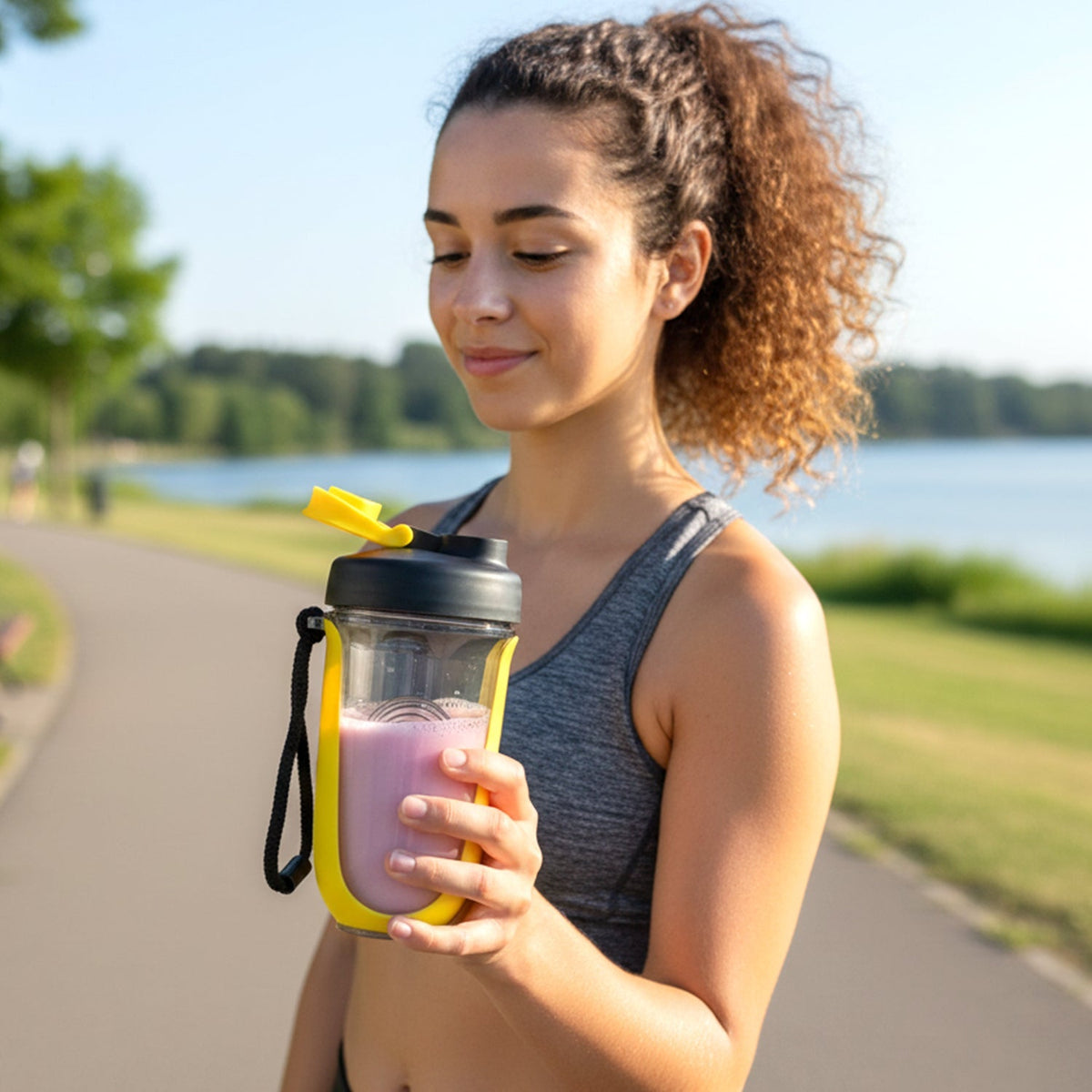 A woman in a grey sports bra holds a yellow and black shaker bottle with pink liquid outdoors by a lake.