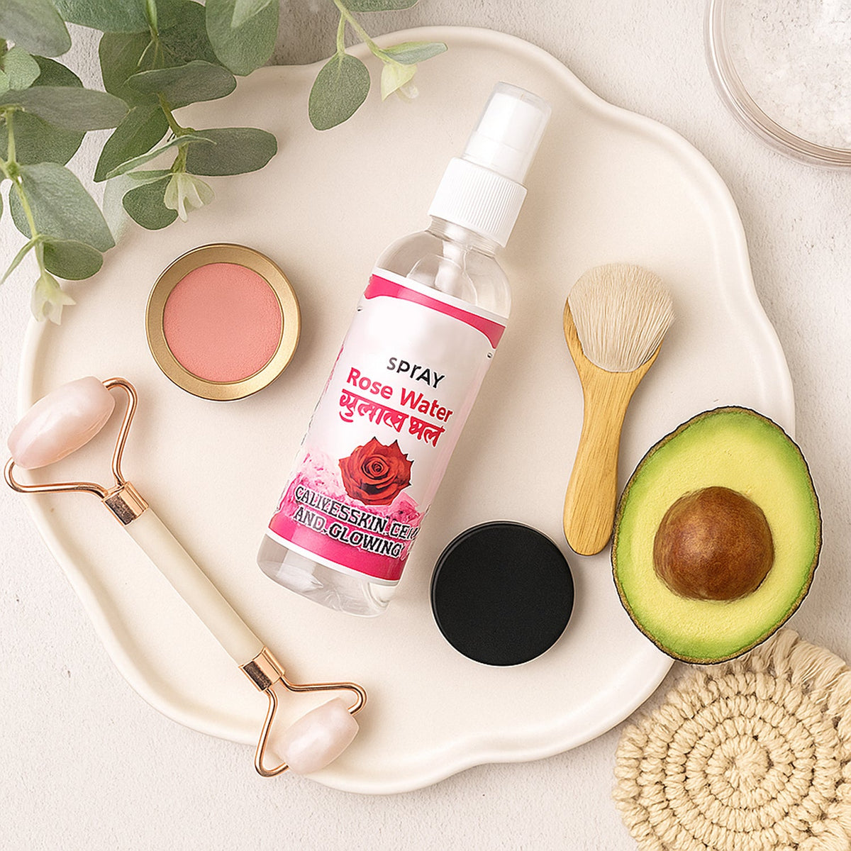 A rose water spray bottle surrounded by a jade roller, blush, brush, avocado, and a coaster on a white tray.