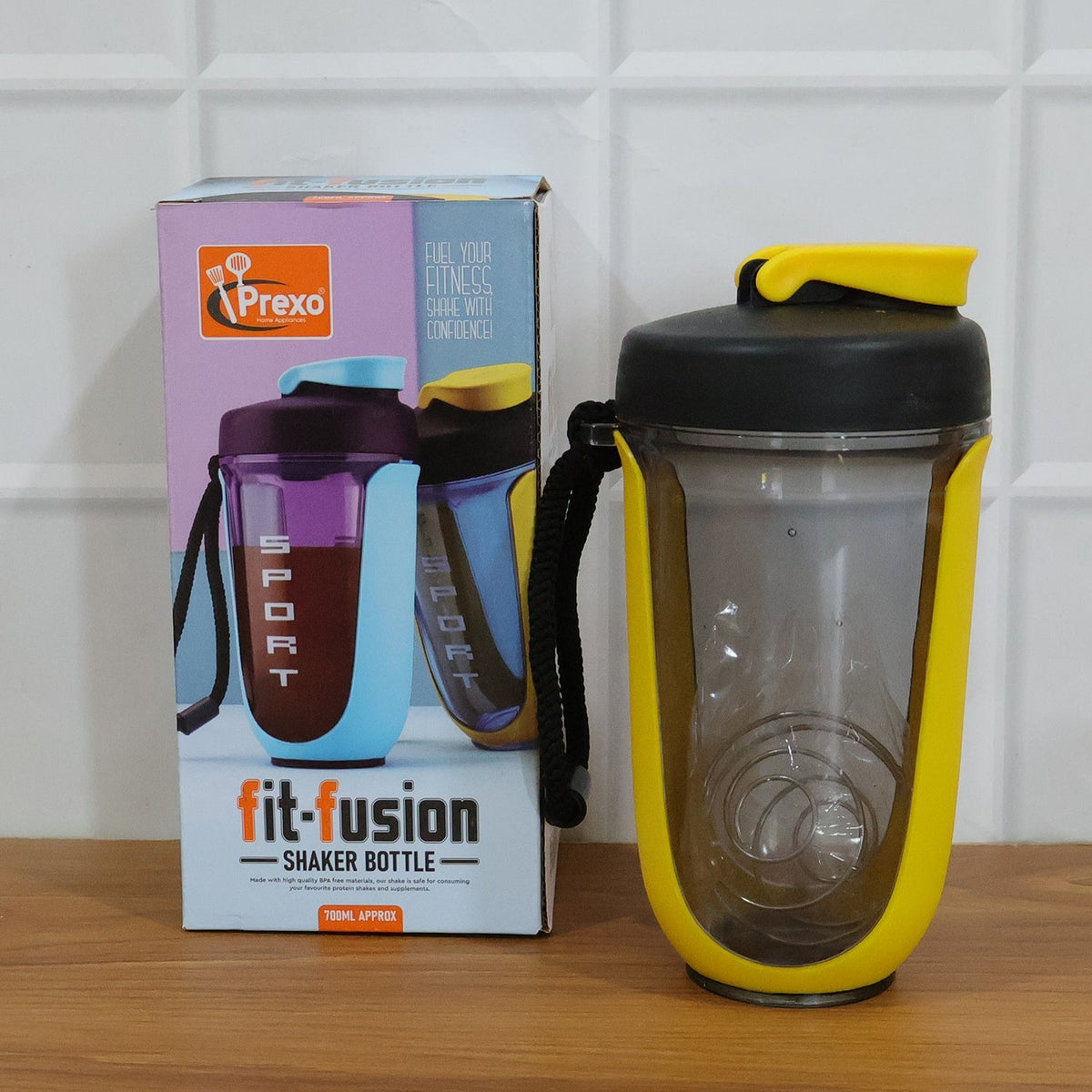A yellow and black shaker bottle with a metal whisk stands next to its colorful product box on a wooden surface.
