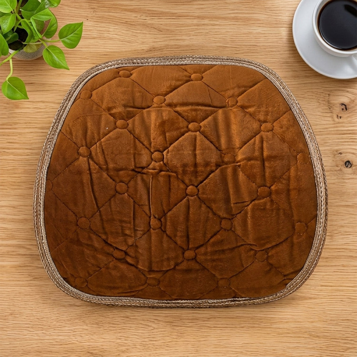 Overhead view of a brown quilted velvet cushion with braided trim on a wooden table.