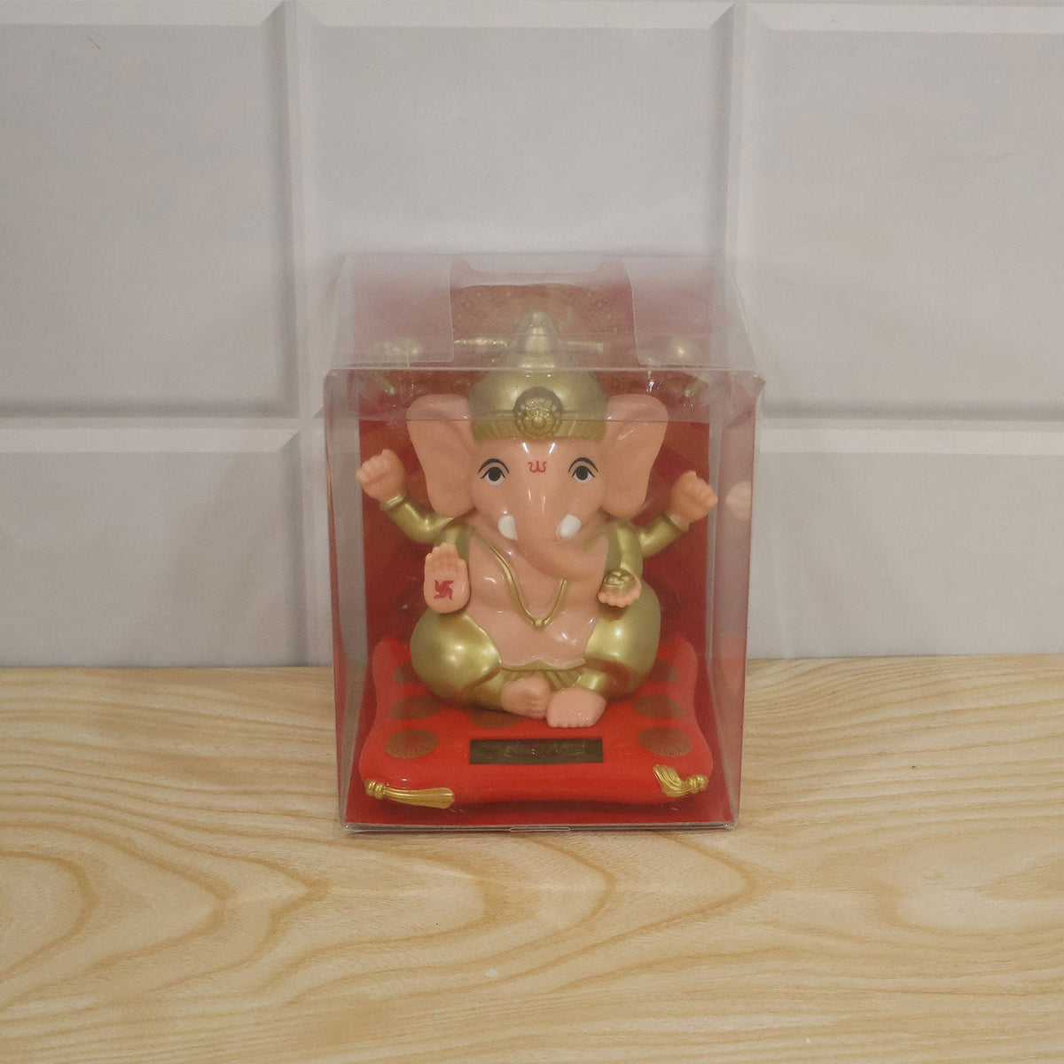 A peach and gold Ganesha idol on a red cushion with a solar panel, packaged in a clear plastic box.