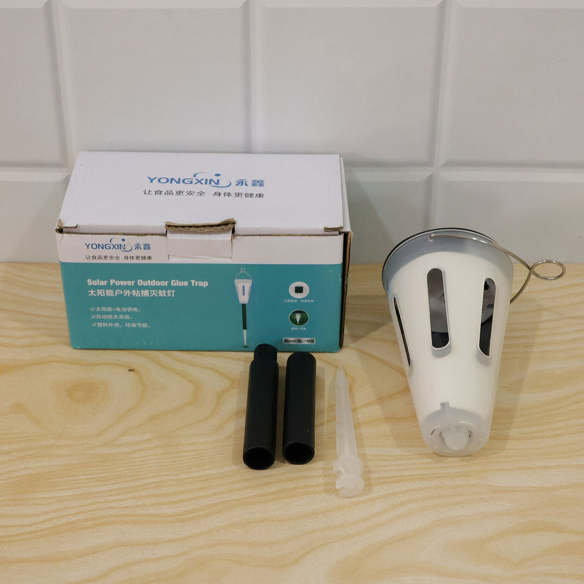 A white box with blue text, a white lantern, two black pole pieces, and a clear stake are on a wooden surface.