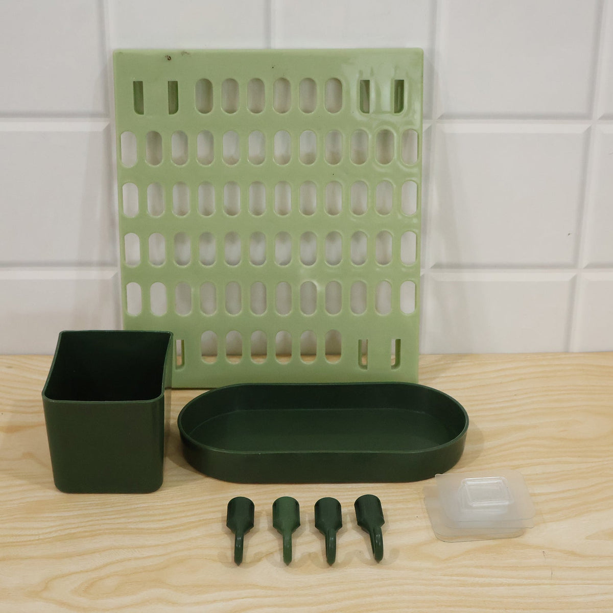 A light green pegboard, a dark green square cup, an oval tray, four hooks, and a clear adhesive pad on a wooden surface with