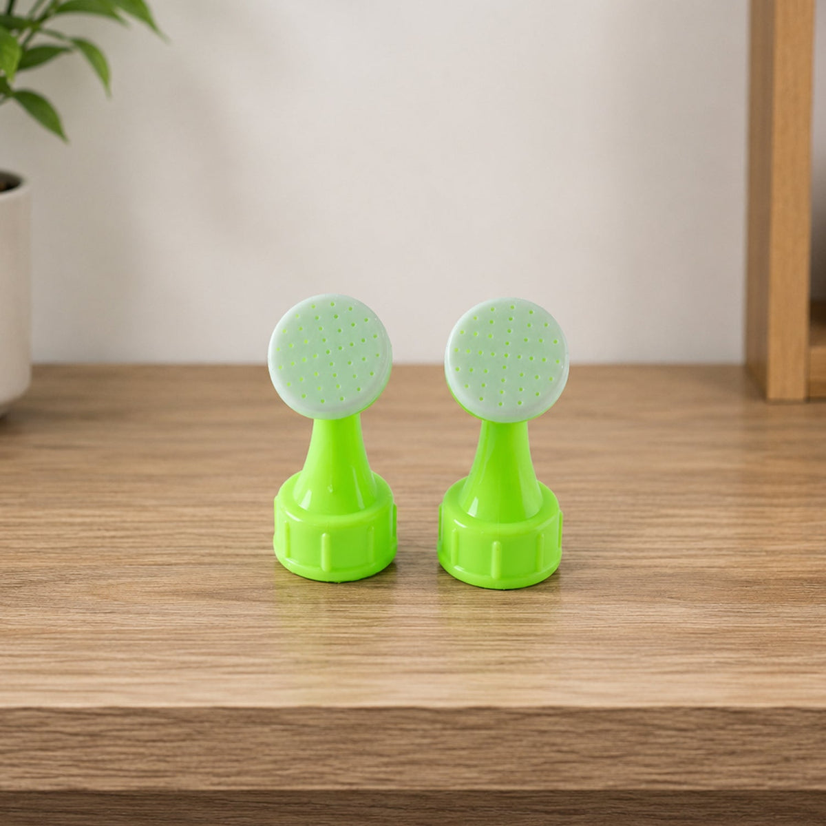 Two identical green plastic bottle top watering sprinklers with white tops are displayed side-by-side on a wooden surface.