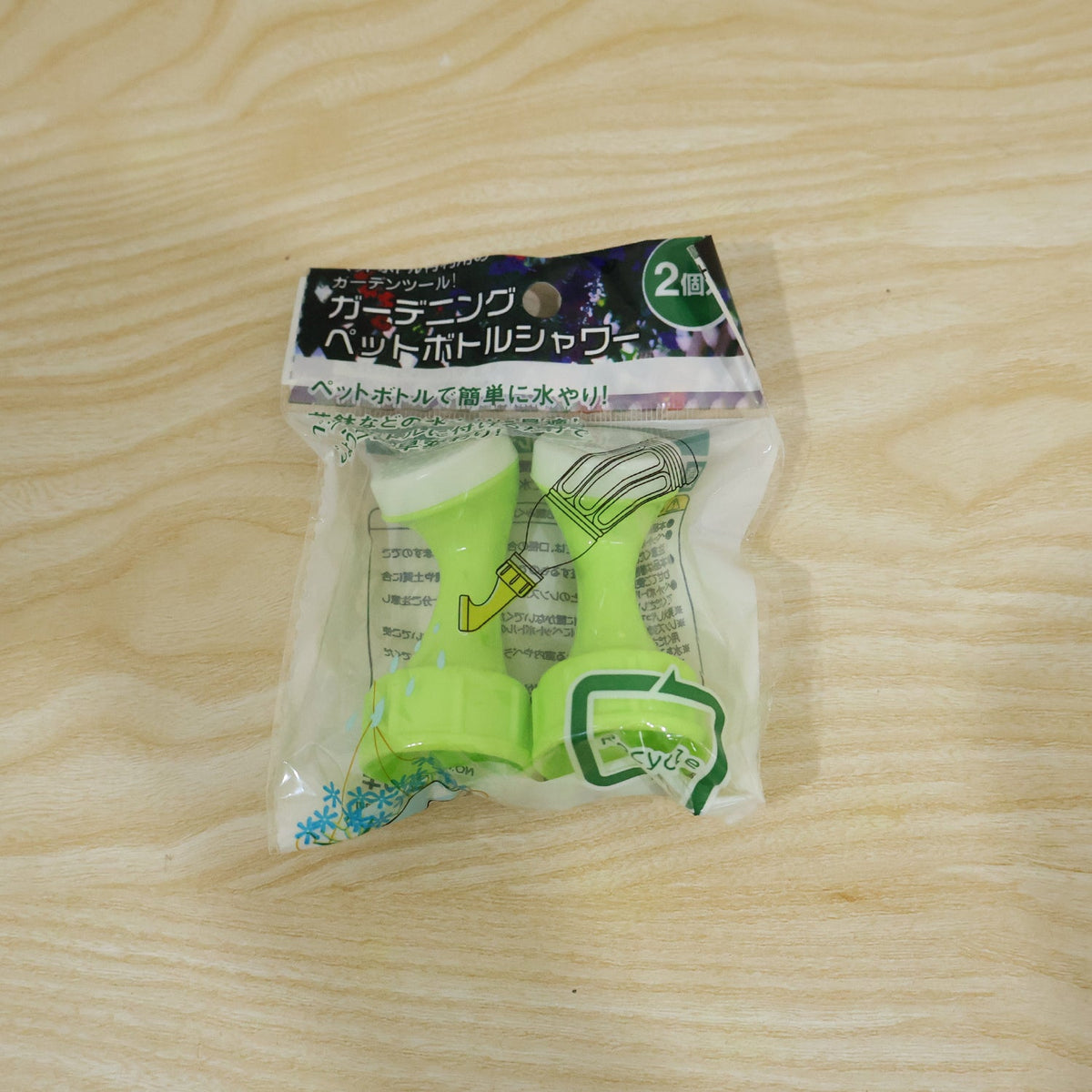 A clear plastic package containing two green plastic bottle top watering sprinklers with Japanese text is on a wooden