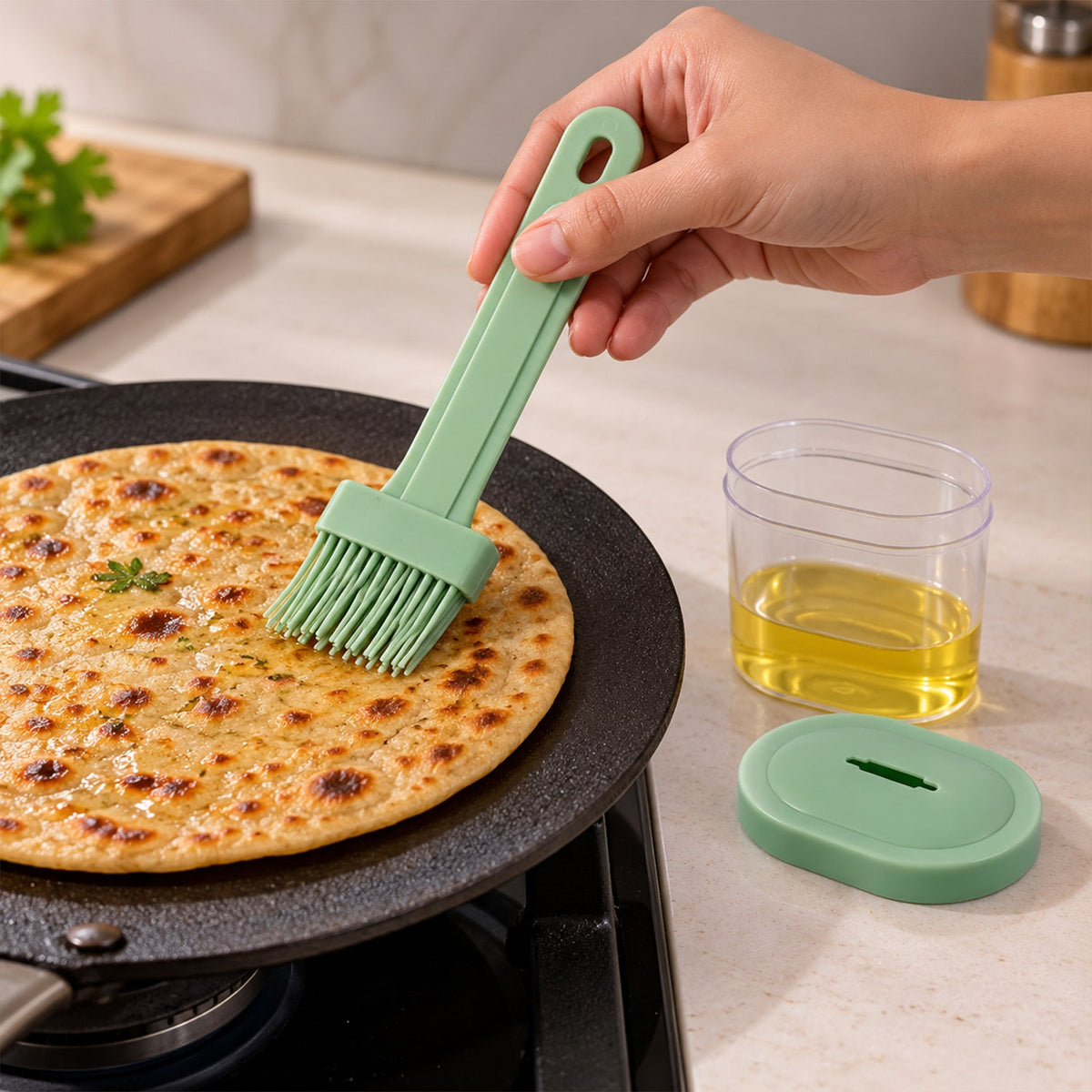 Hand holding a green silicone brush applying oil to a round flatbread cooking on a black griddle. A clear container with oil