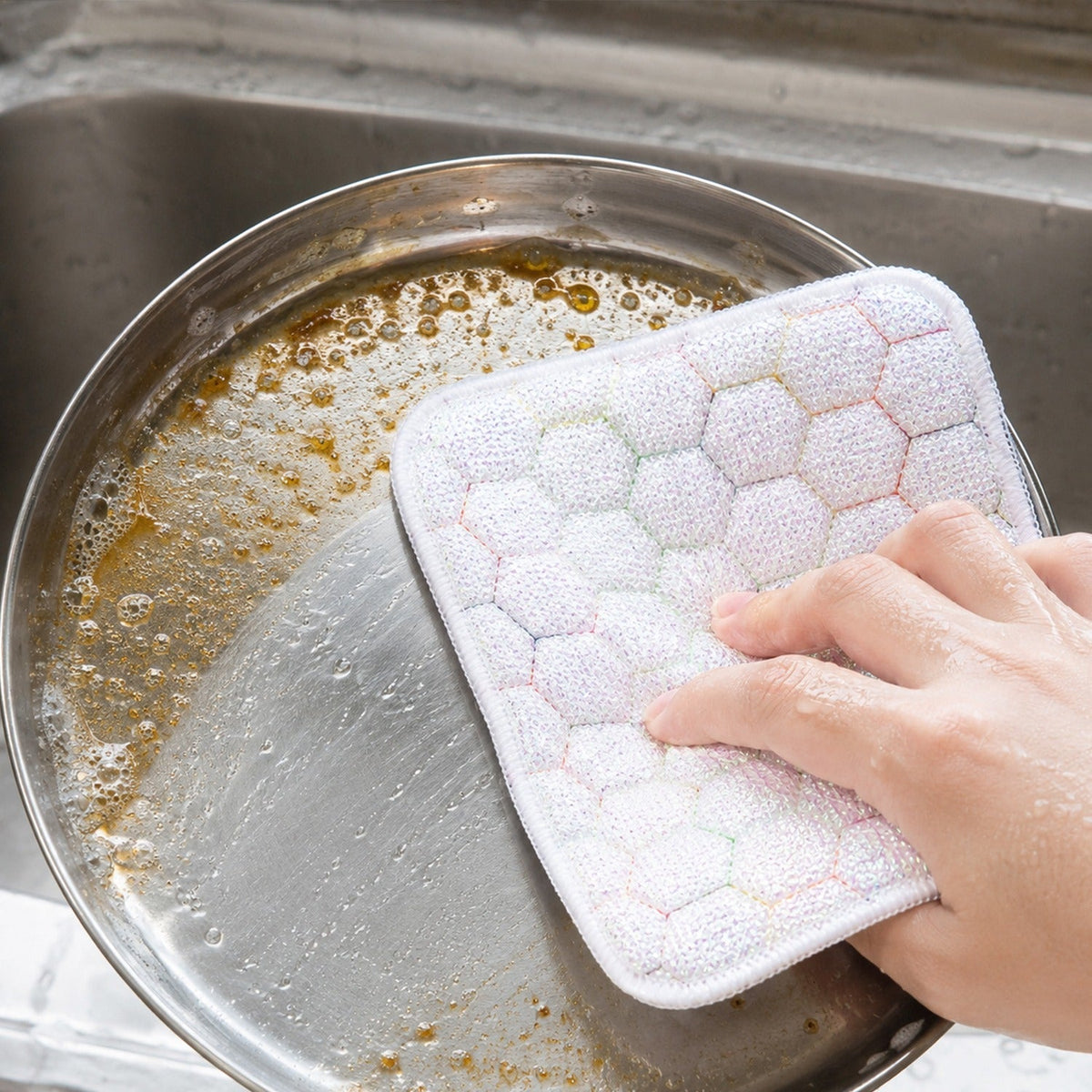 Hand scrubbing a dirty pan with a white honeycomb-patterned cleaning cloth, creating suds in a kitchen sink.