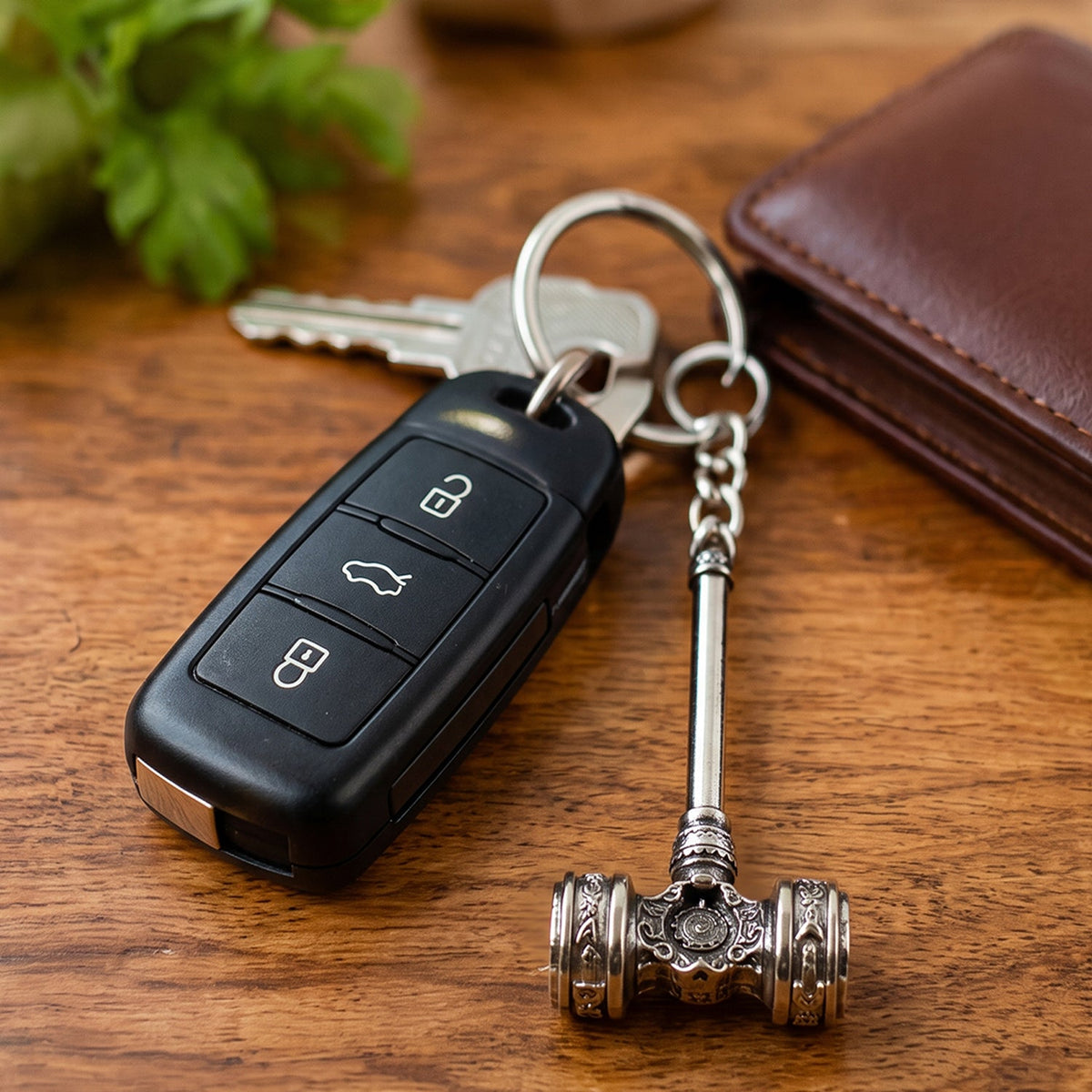 Silver-plated hammer keychain with a car remote and house keys, next to a brown wallet on a wooden surface.