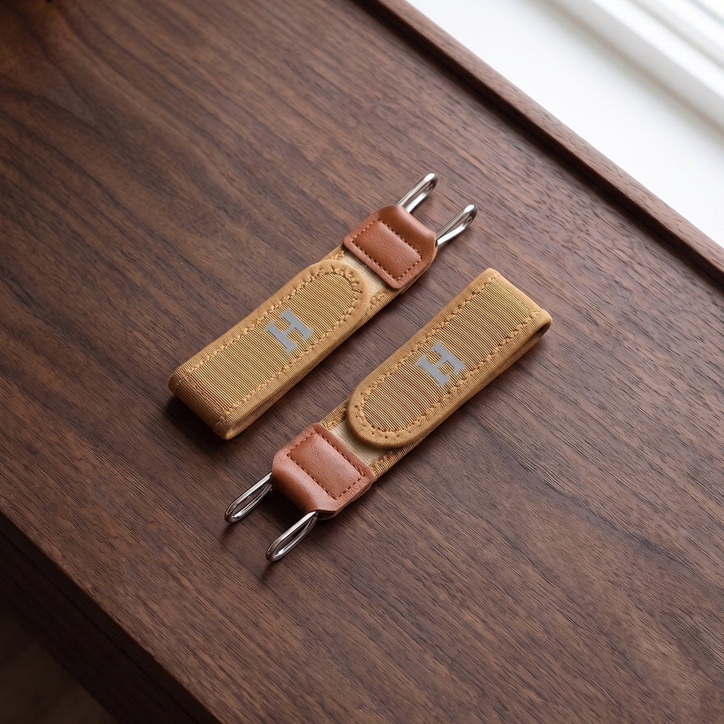 Two tan elastic belt hooks with brown faux leather accents and silver-colored metal hooks, resting on a dark wooden surface.
