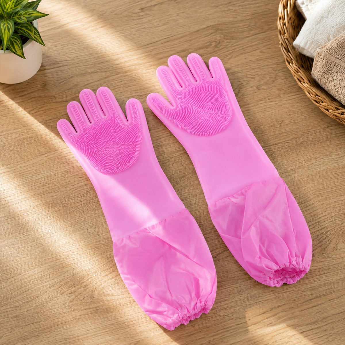 A pair of pink silicone scrubbing gloves with long cuffs on a wooden table next to a plant and a basket.