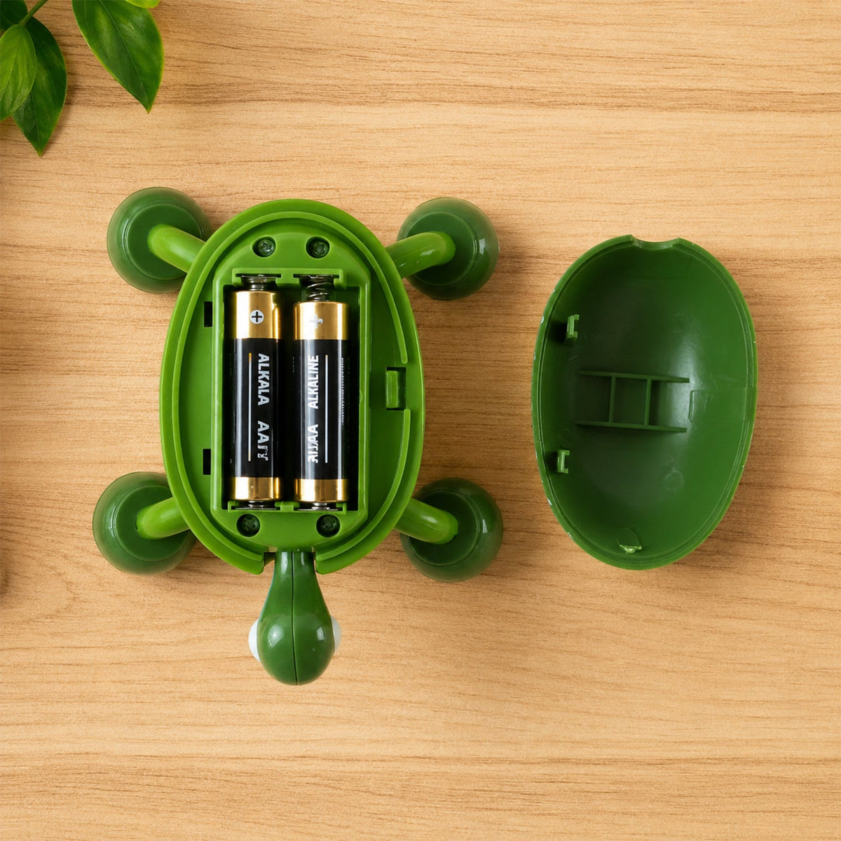 The green turtle massager is open, revealing two AA batteries inside, resting on a wooden surface next to its shell.