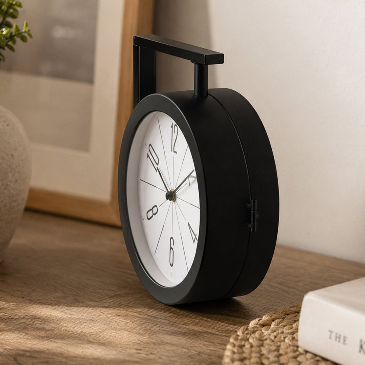 A black double-sided clock with a white face and black numbers sits on a light brown wooden desk next to a book and a plant.