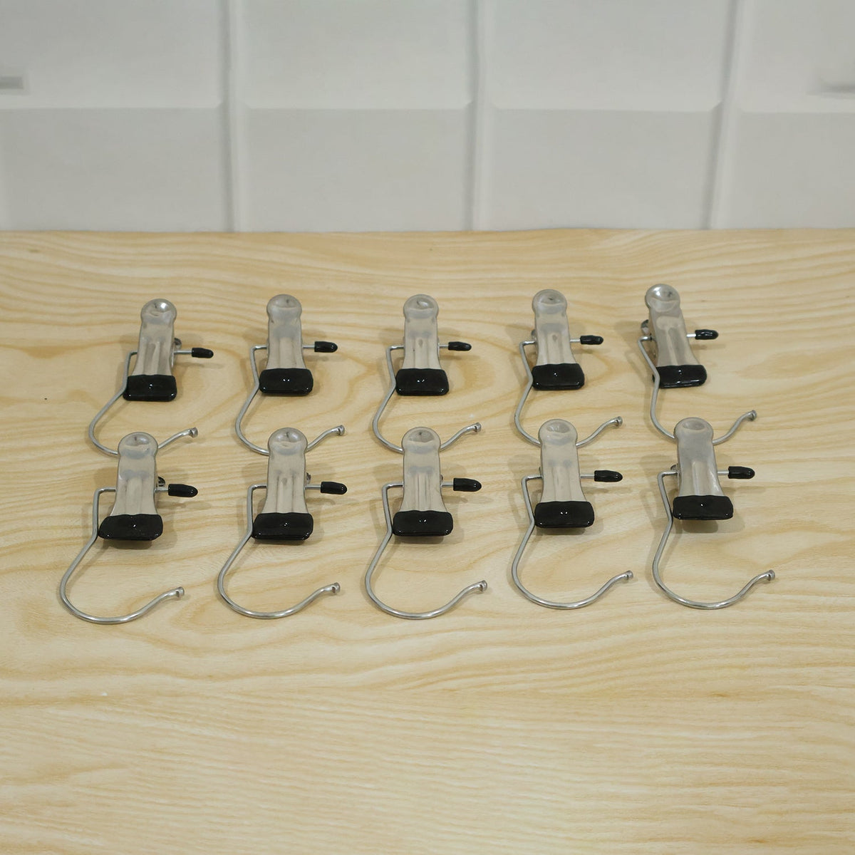 Ten metal clip hangers with black rubber tips and S-shaped hooks are arranged in two rows on a light wooden surface.