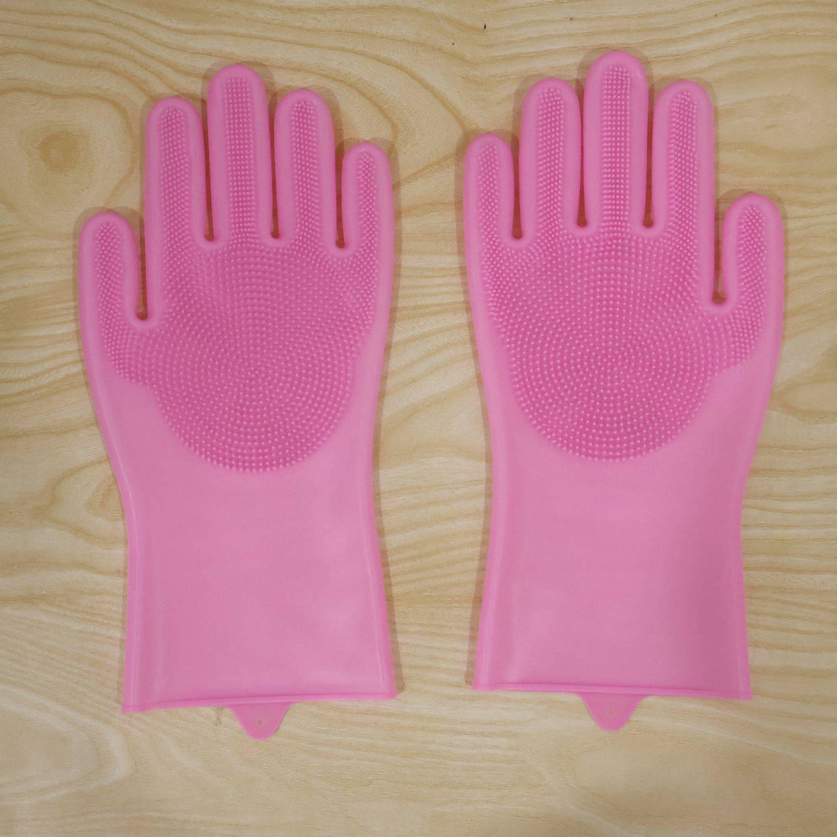 A pair of pink silicone scrubbing gloves with textured palms laid flat on a light wooden surface.