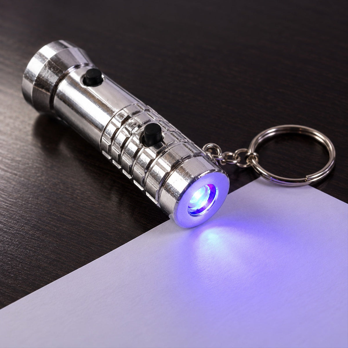 A silver keychain flashlight with a keyring emits a purple UV light onto a white surface on a dark background.
