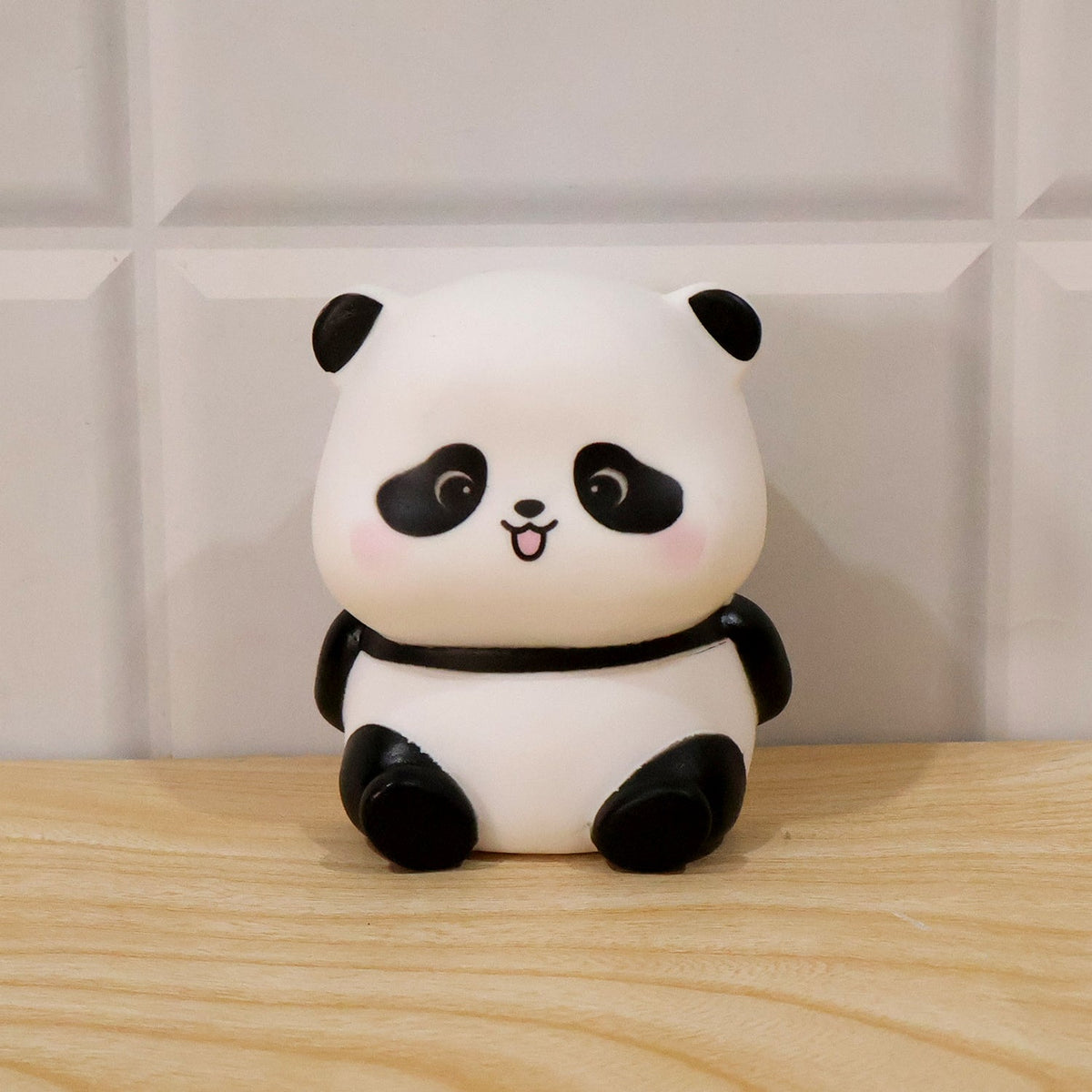 A panda night light, with black ears, eye patches, and limbs, sits on a wooden surface against a white tiled wall.