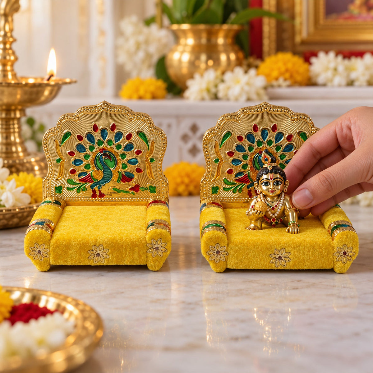 A hand places a small golden idol onto a yellow velvet singhasan with a detailed peacock backrest, on a white surface.