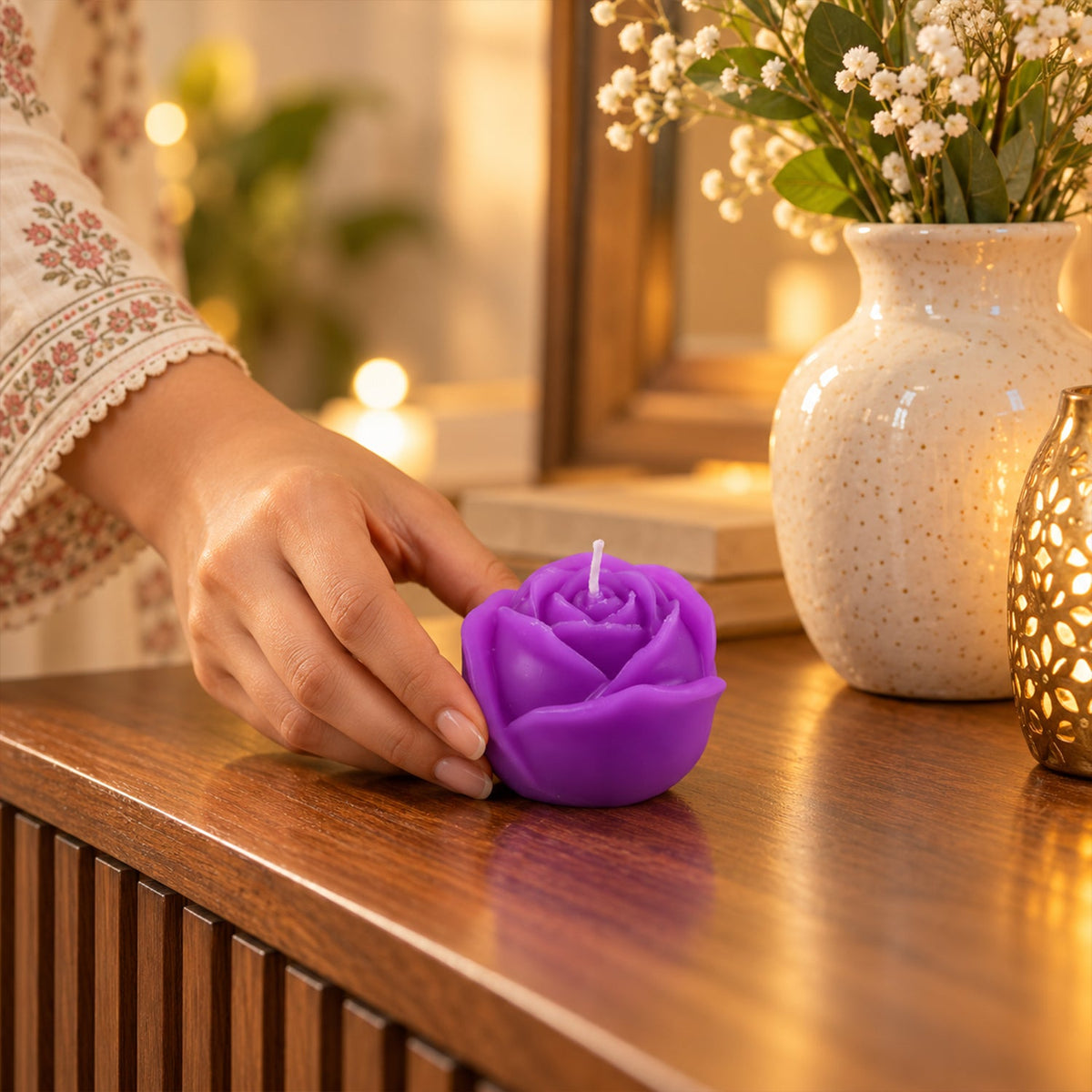 A hand gently places a purple rose-shaped candle on a ribbed wooden dresser next to a white vase with flowers.