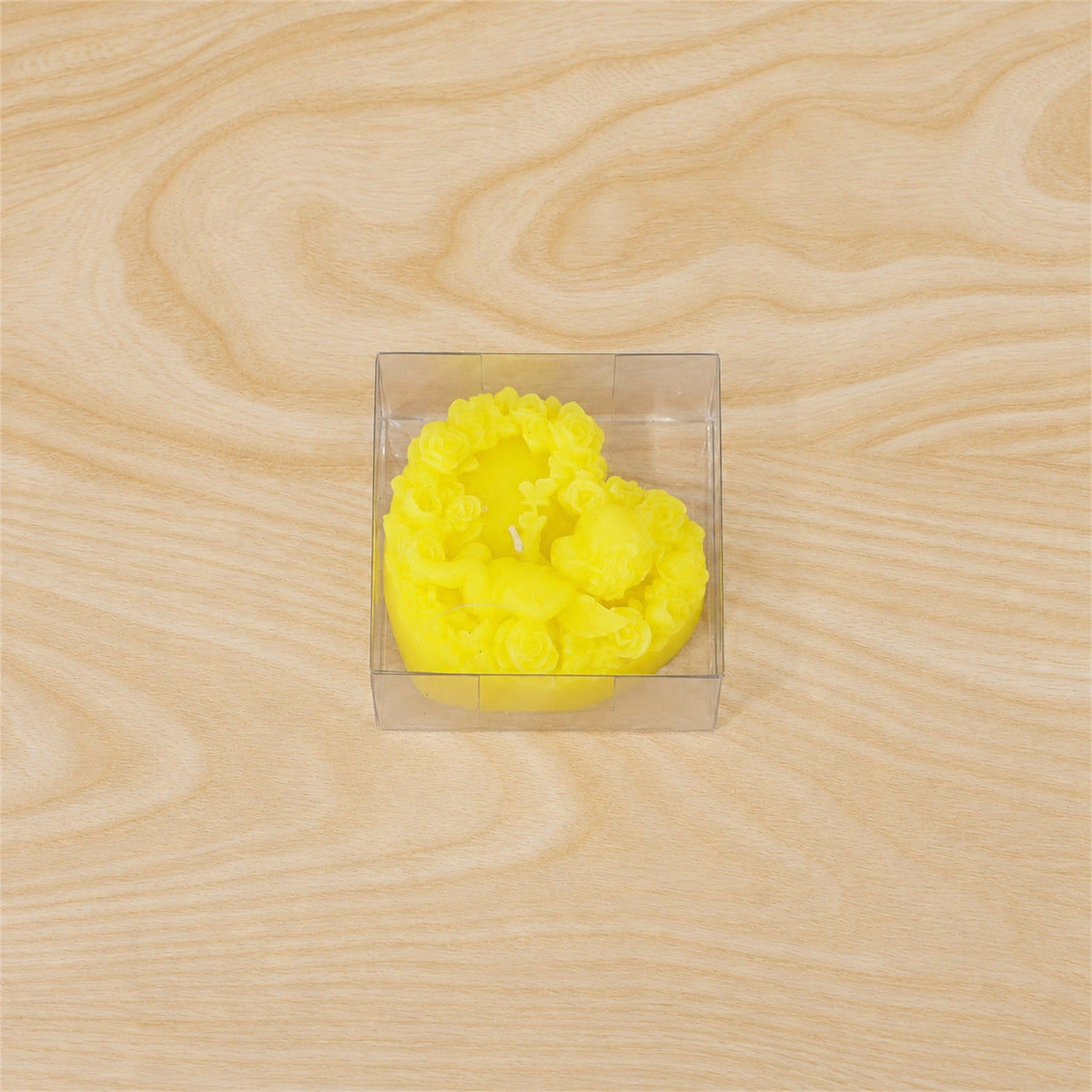 A yellow heart-shaped candle with a cherub design, neatly packaged in a clear square box on a wooden surface.