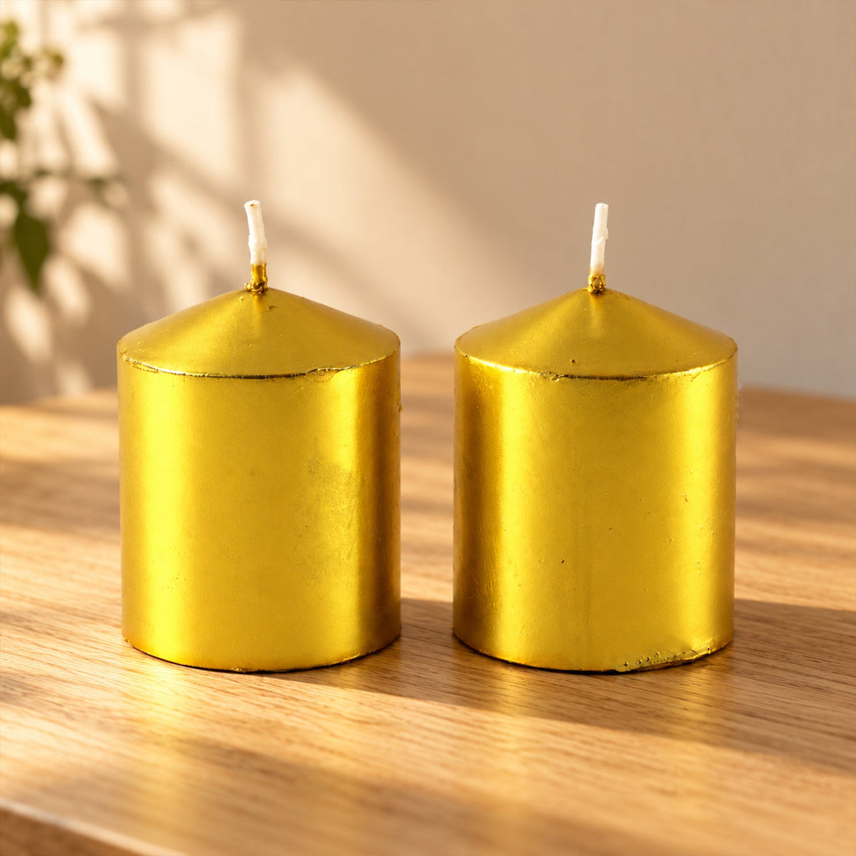 Two unlit golden metallic pillar candles with white wicks on a light wooden surface in natural sunlight.