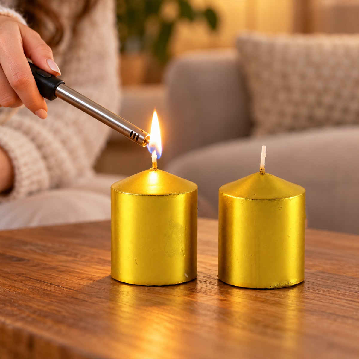 A person's hand lighting one of two golden metallic pillar candles on a dark wooden table.