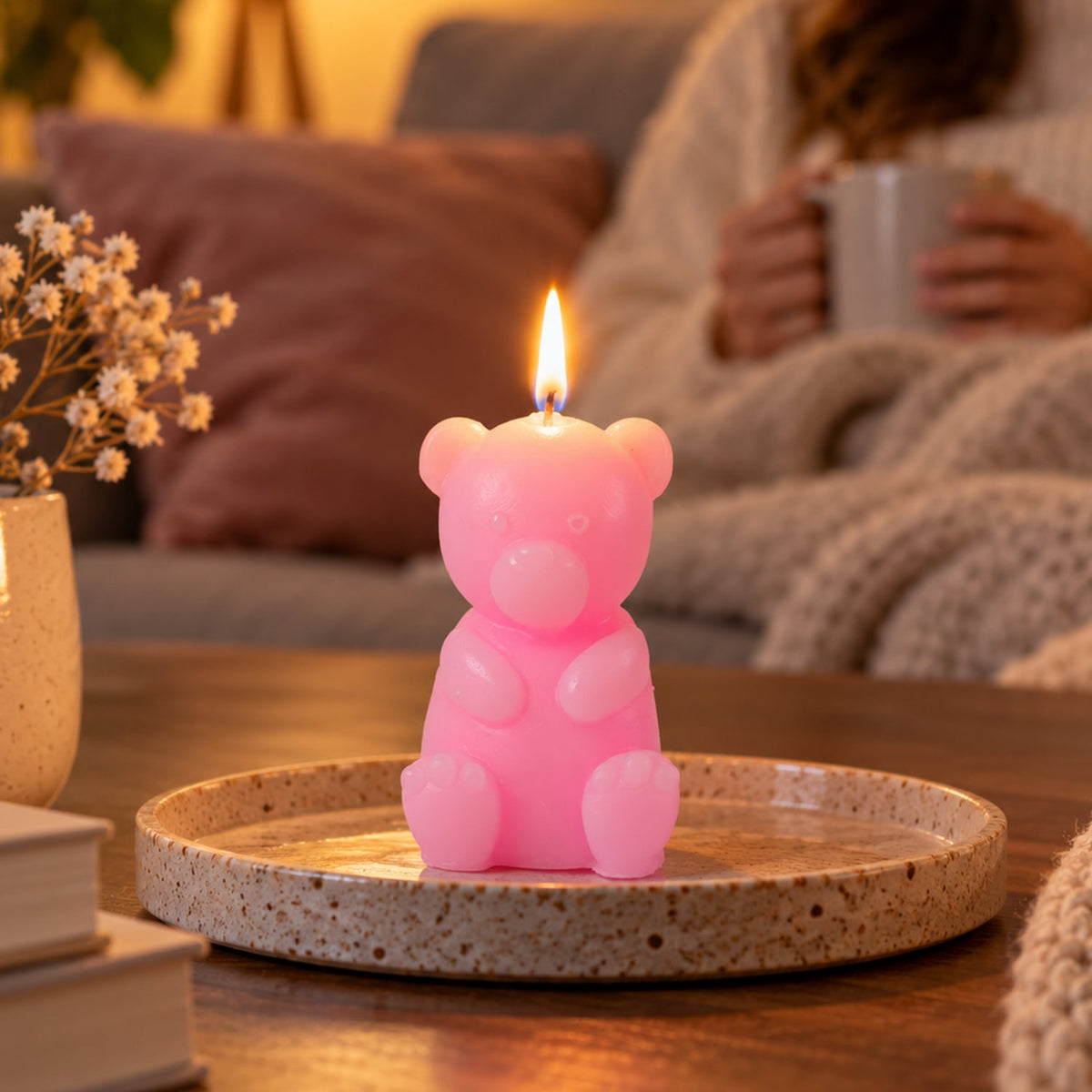 A lit pink teddy bear candle sits on a speckled ceramic plate, with a person holding a mug in the background.