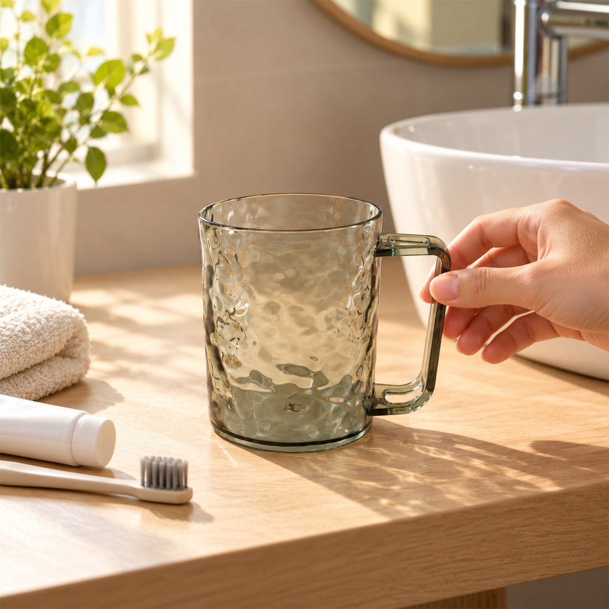 A hand reaching for a dark grey textured plastic mug on a wooden bathroom counter next to a white sink.