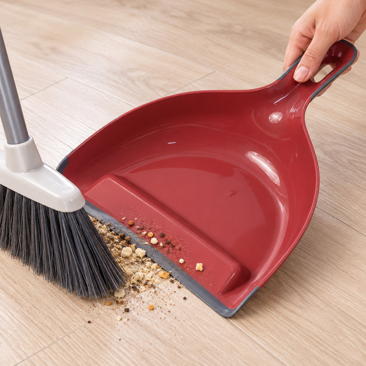 A person sweeps debris into a red and grey dustpan using a grey broom on a light wooden floor.