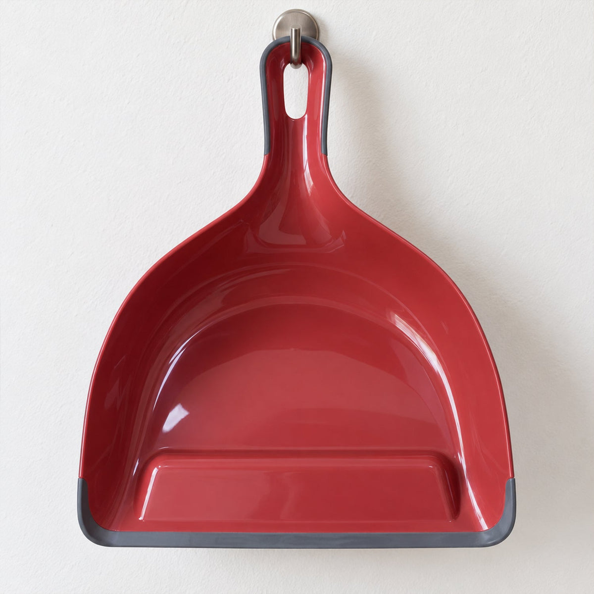 A red and grey dustpan hangs from a silver hook on a plain white wall.