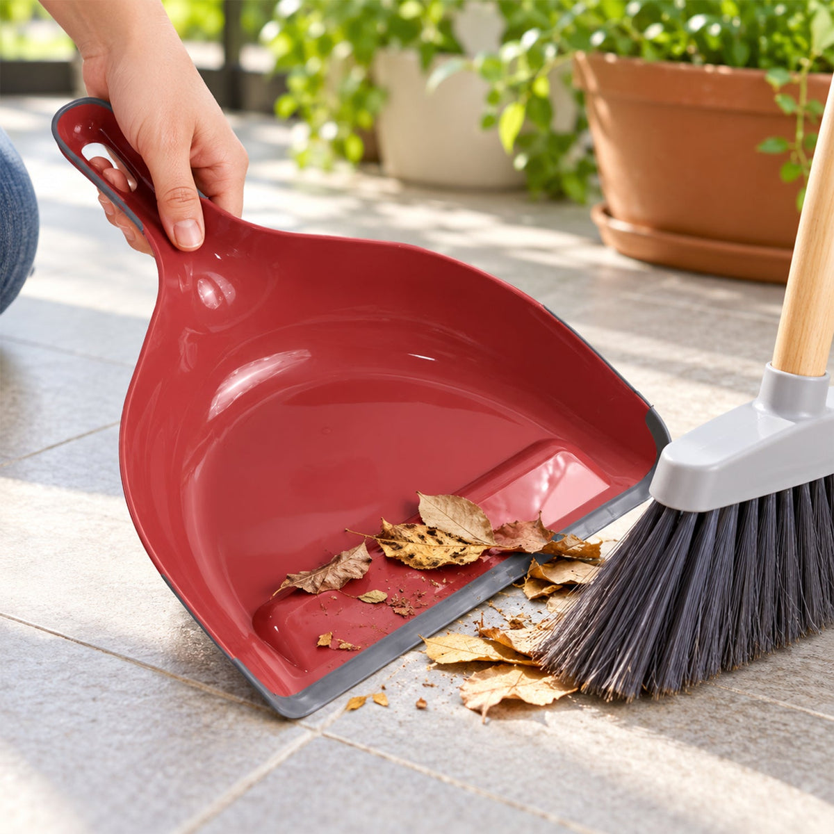 A person sweeps dry leaves into a red and grey dustpan on an outdoor patio with potted plants.