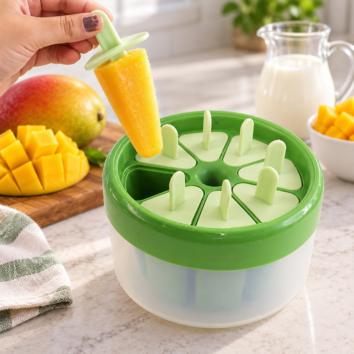 A hand removes a yellow mango ice cream from a green and white round plastic mold, with fresh mango and milk nearby.