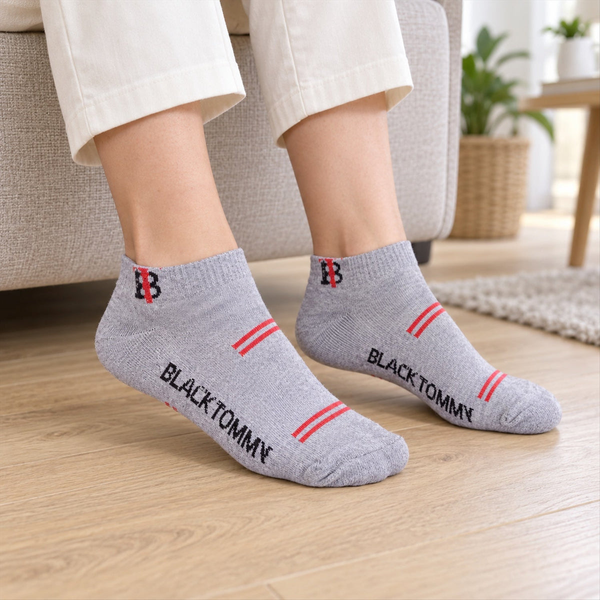 A person wears light grey ankle socks with red stripes and "BLACK TOMMY" text on a wooden floor.