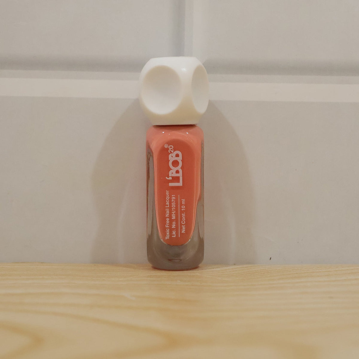 A single bottle of peach coral nail polish with a white dice-shaped cap stands on a light wooden surface against a white