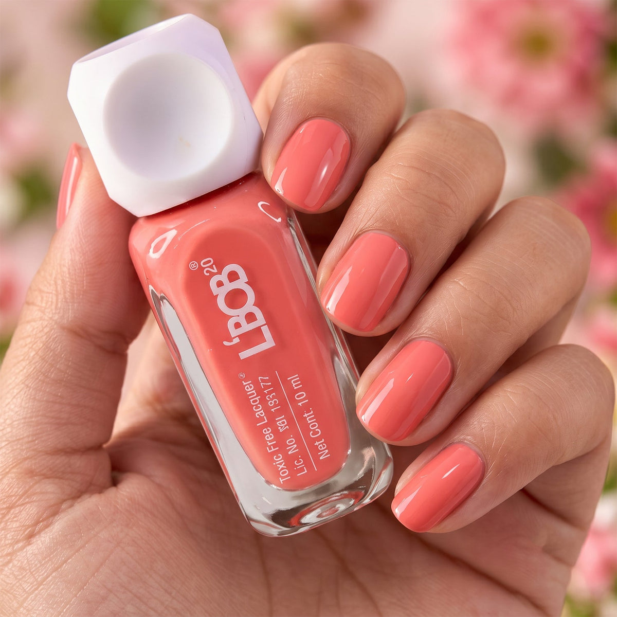 A hand holds a bottle of peach coral nail polish, with the fingernails painted in the same vibrant shade against a blurred