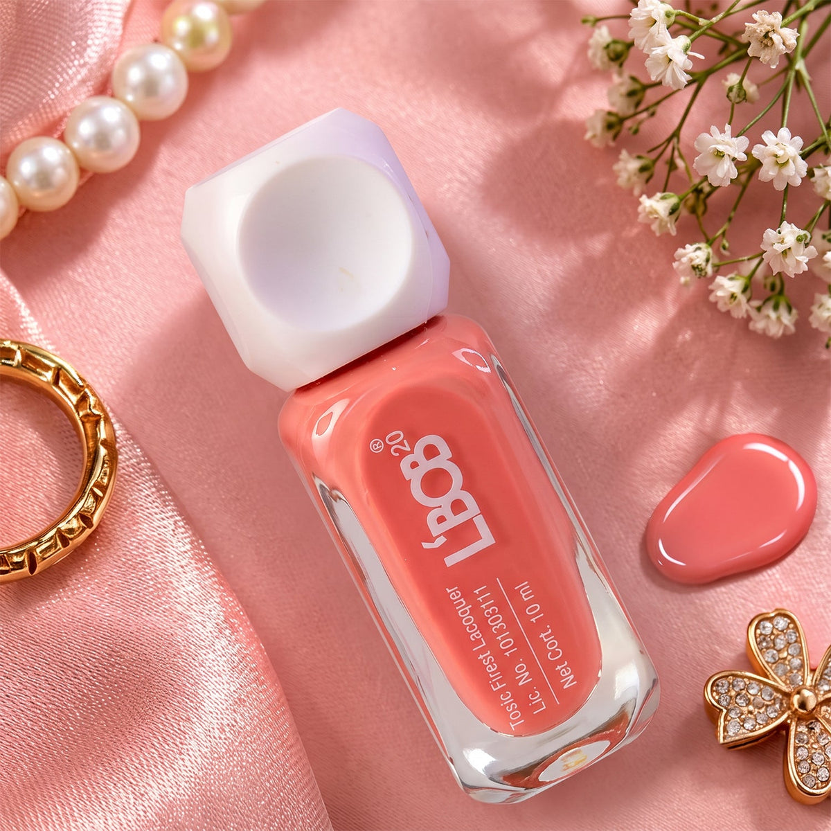 A peach coral nail polish bottle rests on a pink satin fabric, surrounded by a string of pearls, a gold ring, white baby's
