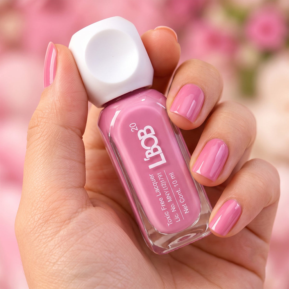 A hand with neatly painted pink nails holds a matching nail polish bottle against a soft pink floral background.