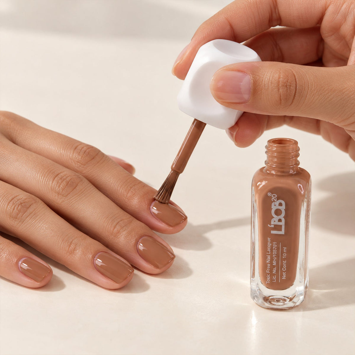 A person applies nude brown polish to fingernails with a brush, next to an open bottle on a white surface.