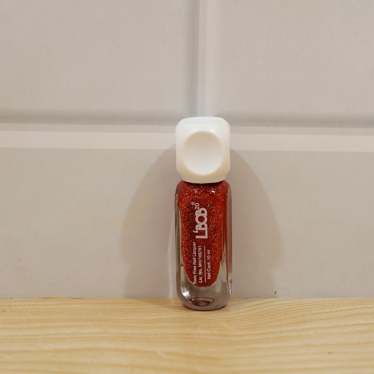 A small, clear glass bottle of red glitter nail polish with a white cap stands on a wooden surface.