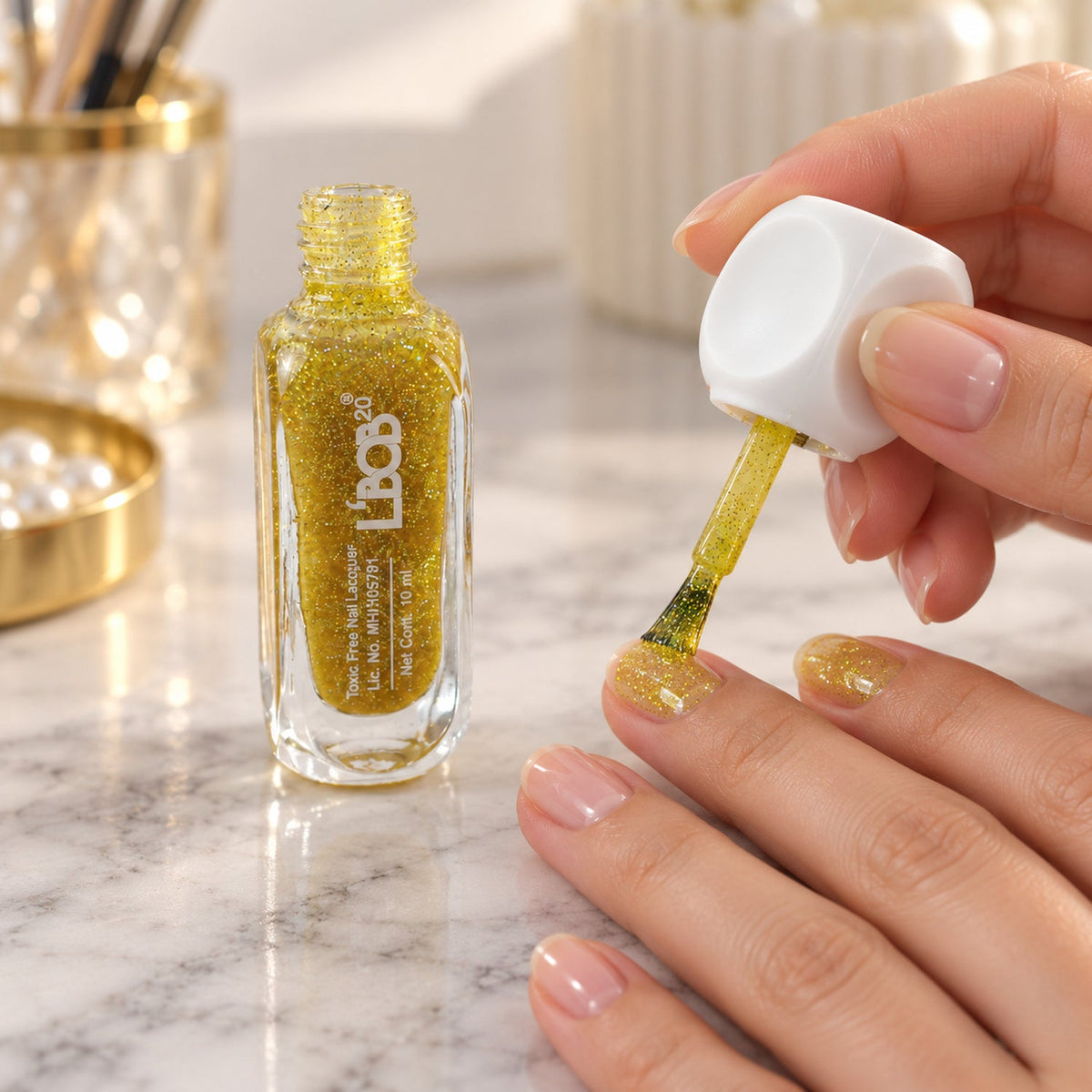 A person applies gold glitter nail polish to their fingernails, with a bottle of polish beside them.
