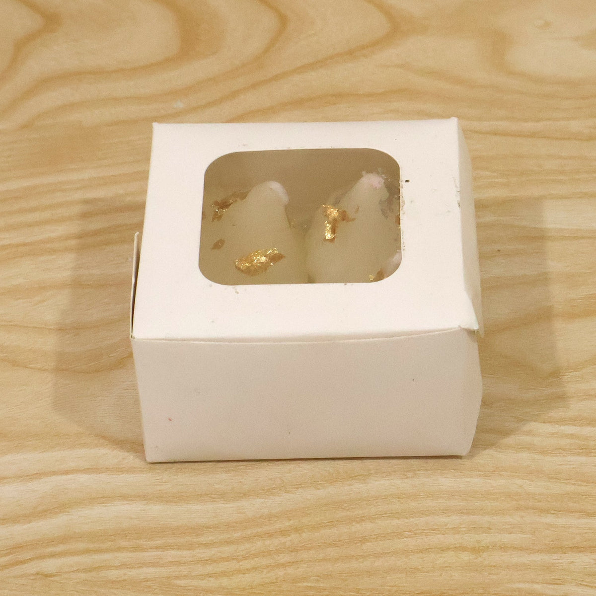 A small white cardboard box with a clear window, displaying two modak-shaped white candles with gold flakes inside.