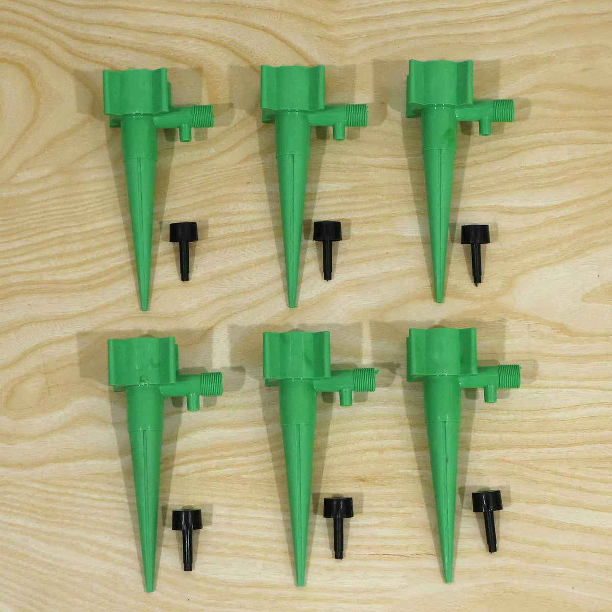Six green plastic plant watering spikes and six small black caps are arranged on a light wooden surface.