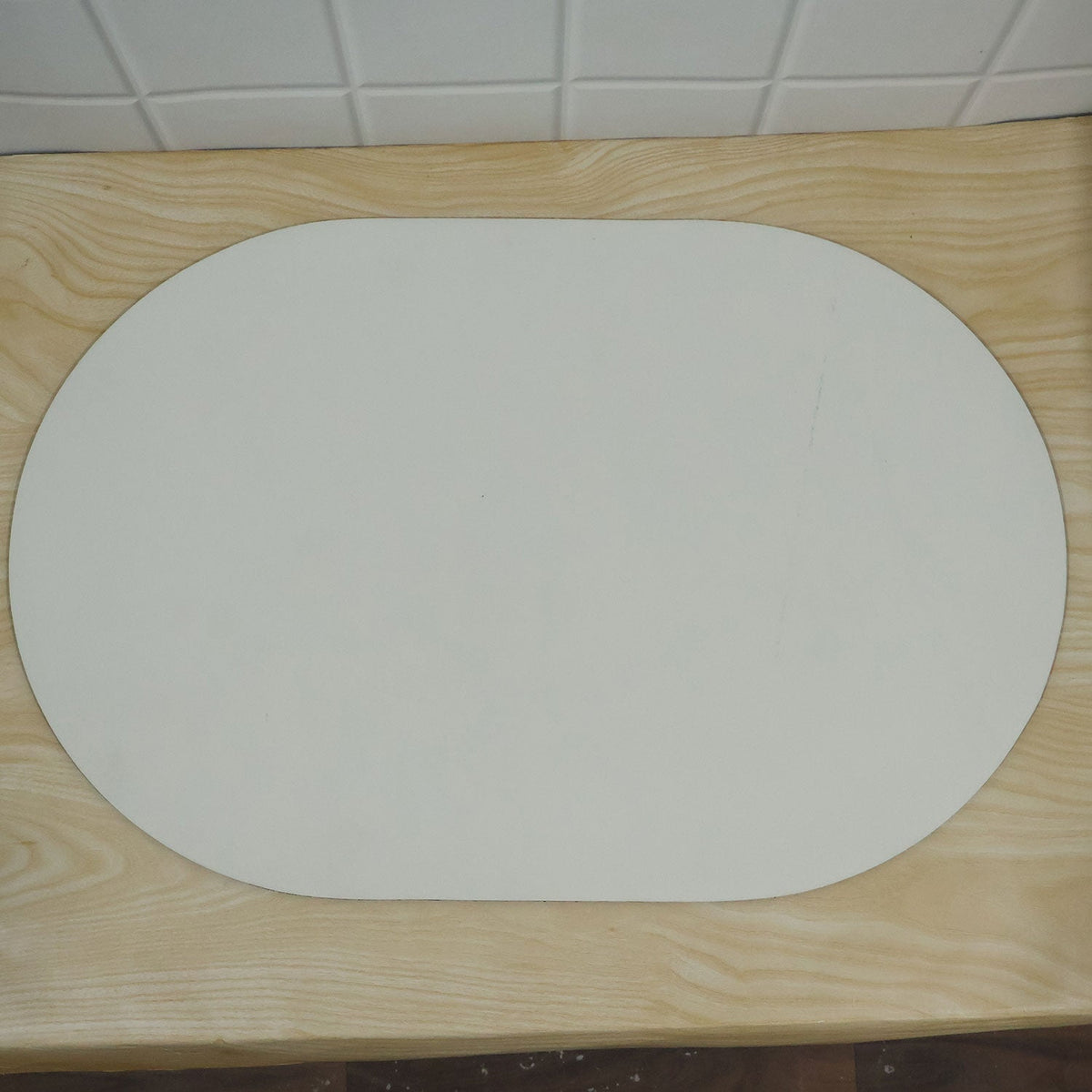 A plain white oval mat resting on a light wooden surface with white tiled background.