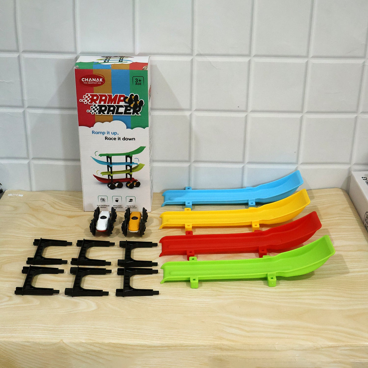 All components of the ramp racer toy set, including the box, ramps, cars, and connectors, arranged on a wooden surface.