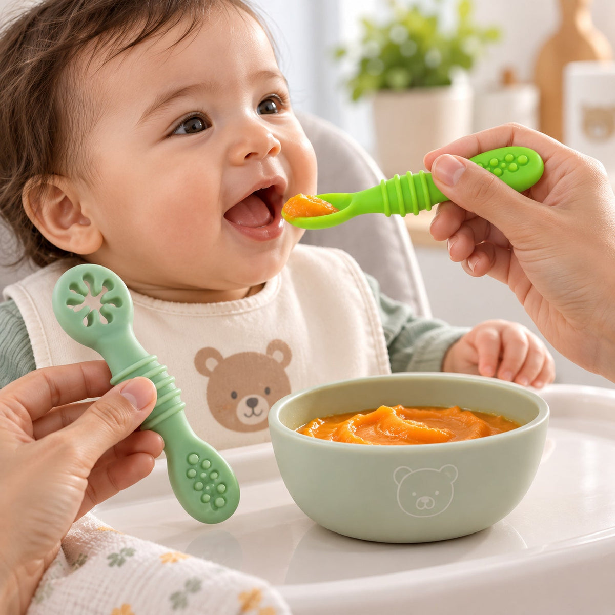 A baby with an open mouth being fed pureed food with a green silicone spoon, with another spoon and a bowl on the high chair